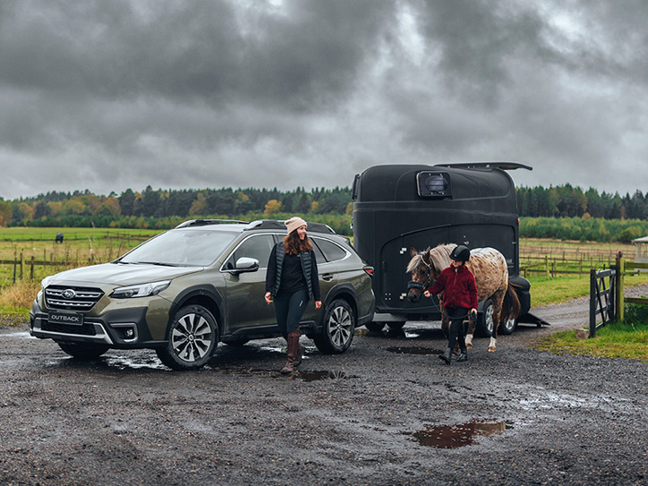 Subaru Outback mudassa hevostrailerin kanssa, samalla kun nainen ja tyttö kävelevät vieressä – tyttö taluttaa hevosta.